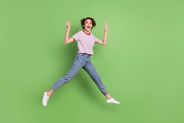 Full length body size view of pretty cheerful amazed girl jumping showing v-sign isolated over green pastel color background - Foto, Bild