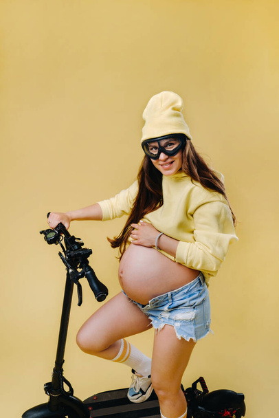 Een zwanger meisje in gele kleren op een elektrische scooter op een geïsoleerde gele achtergrond. - Foto, afbeelding