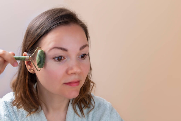 Mooie vrouw die jade face roller vasthoudt en gezichtsmassages maakt. Huidverzorging thuis - Foto, afbeelding