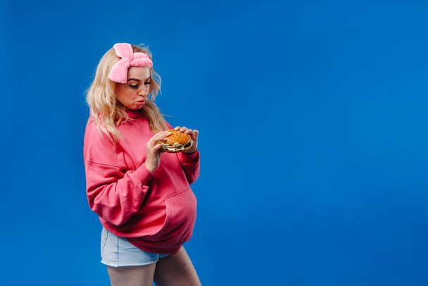 a pregnant girl in pink clothes with a hamburger in her hands on a blue background. - Фото, зображення