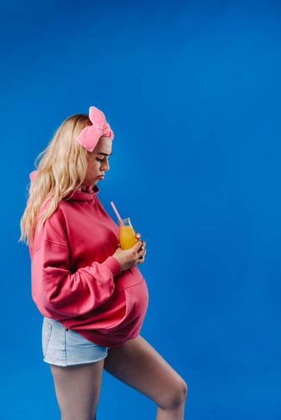 pregnant girl in pink clothes with a bottle of juice on a blue background. - Foto, Imagen