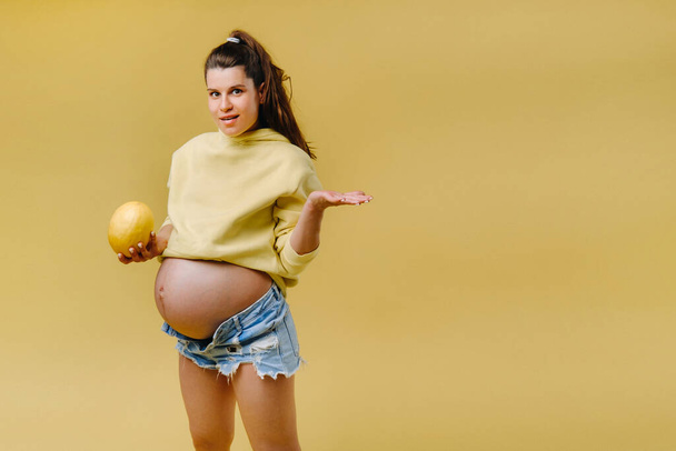 a pregnant girl with a yellow jacket stands with a melon in her hands on a yellow isolated background. - Фото, зображення