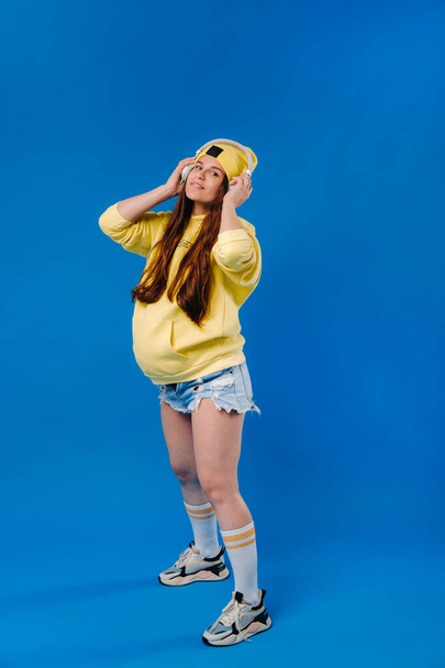 A pregnant girl in a yellow jacket and headphones stands on a blue background. - Foto, afbeelding