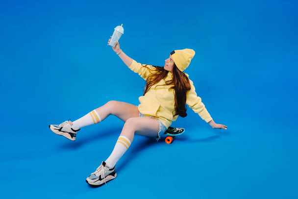 těhotná dívka ve žlutém oblečení se sklenicí šťávy sedí na skateboardu na modrém pozadí. - Fotografie, Obrázek