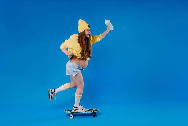 a pregnant girl in yellow clothes with a glass of juice rides a skateboard on a blue background. - Fotoğraf, Görsel