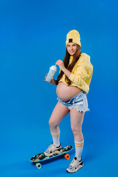 a pregnant girl in yellow clothes with a glass of juice rides a skateboard on a blue background. - Fotoğraf, Görsel