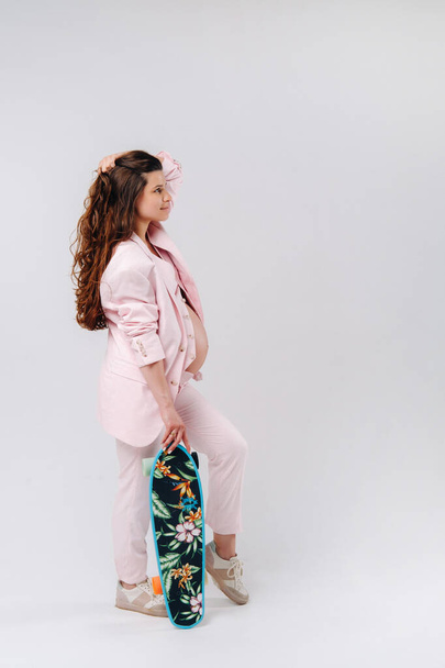 A pregnant girl in a pink suit with a skateboard in her hands stands on a gray background. - Foto, imagen