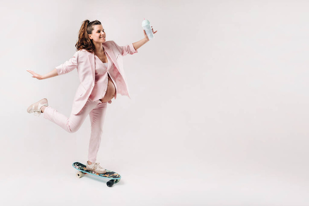 A pregnant girl in a pink suit with a skateboard in her hands stands on a gray background. - Foto, Imagen