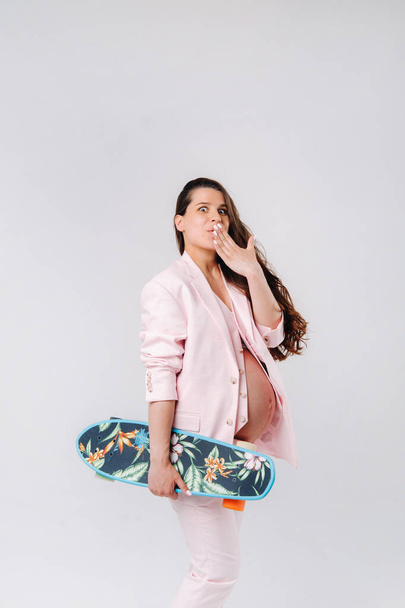 A pregnant girl in a pink suit with a skateboard in her hands stands on a gray background. - Photo, Image