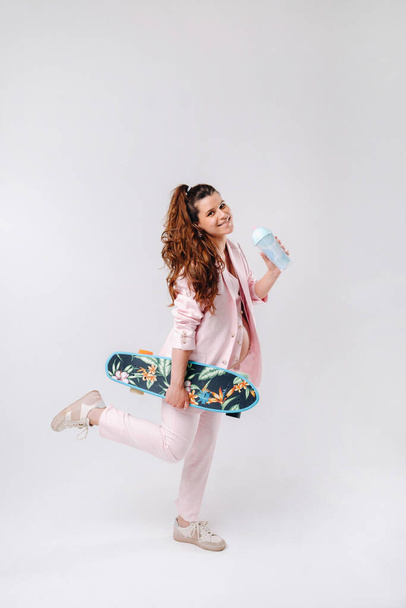 A pregnant girl in a pink suit with a skateboard in her hands drinks juice on a gray background. - Foto, immagini
