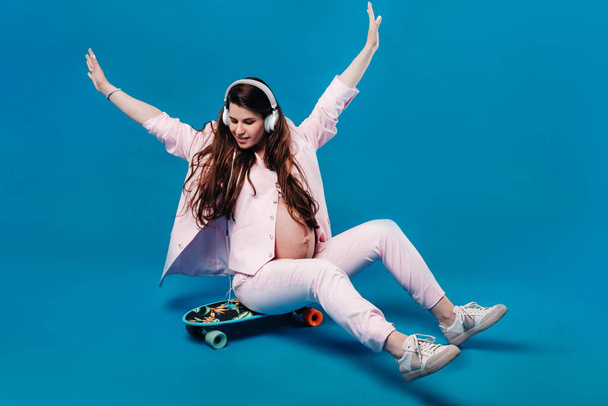 a pregnant woman in a pink suit sits on a skateboard with headphones and listens to music on a blue background. - Photo, image