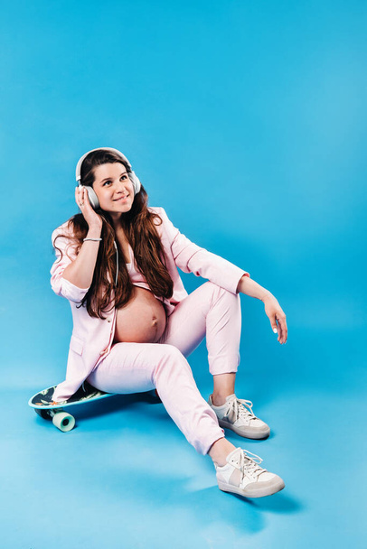 a pregnant woman in a pink suit sits on a skateboard with headphones and listens to music on a blue background. - Photo, Image