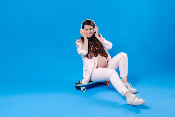 a pregnant woman in a pink suit sits on a skateboard with headphones and listens to music on a blue background. - Zdjęcie, obraz