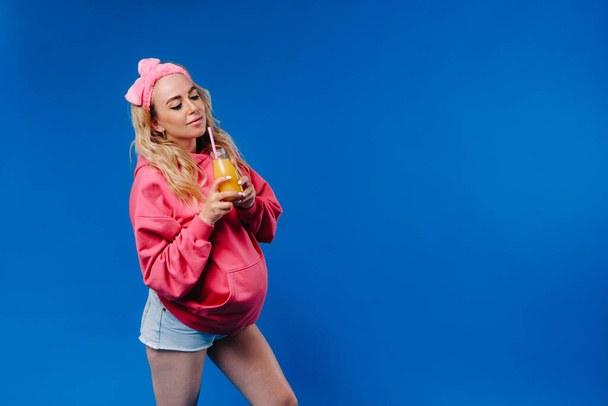 pregnant girl in pink clothes with a bottle of juice on a blue background. - Valokuva, kuva
