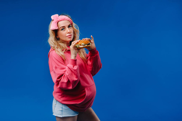 a pregnant girl in pink clothes with a hamburger in her hands on a blue background. - Photo, Image