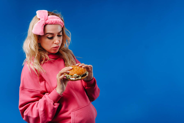 a pregnant girl in pink clothes with a hamburger in her hands on a blue background. - Valokuva, kuva