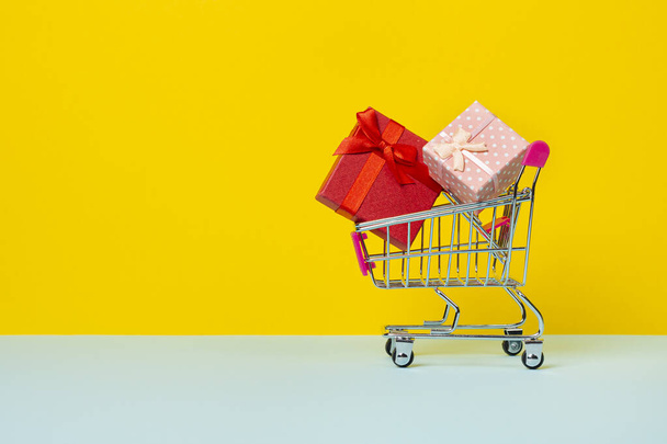 Toy shopping cart or trolley with full with gift boxes. A gift for a holiday, anniversary or birthday. Trolley cart with gift boxes Isolated over yellow background - Photo, Image