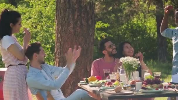 Grupo de hombres y mujeres modernos y alegres que se divierten en la fiesta en el parque almorzando, escuchando música y bailando en un día soleado - Imágenes, Vídeo