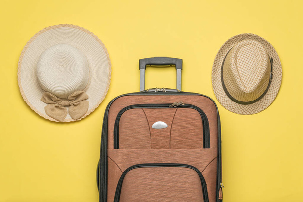 Ανδρικά και γυναικεία καπέλα και μια βαλίτσα ταξιδιού σε κίτρινο φόντο. Η έννοια του καλοκαιρινού ταξιδιού. Επίπεδη. - Φωτογραφία, εικόνα