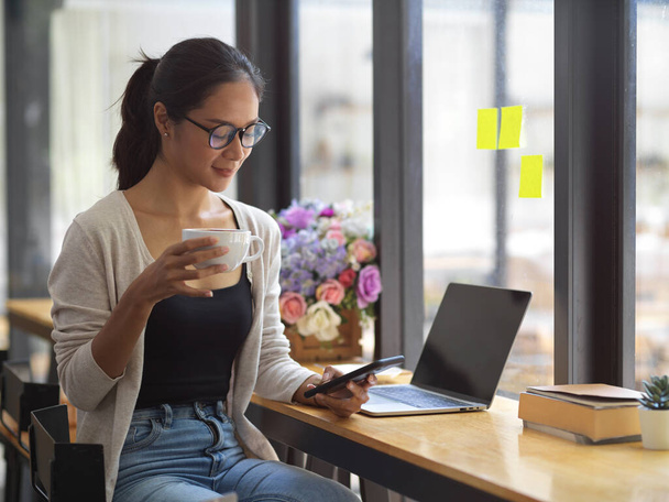 Atraktivní žena na volné noze s brýlemi k pití kávy a rolování na smartphonu s otevřeným notebookem v kavárně - Fotografie, Obrázek