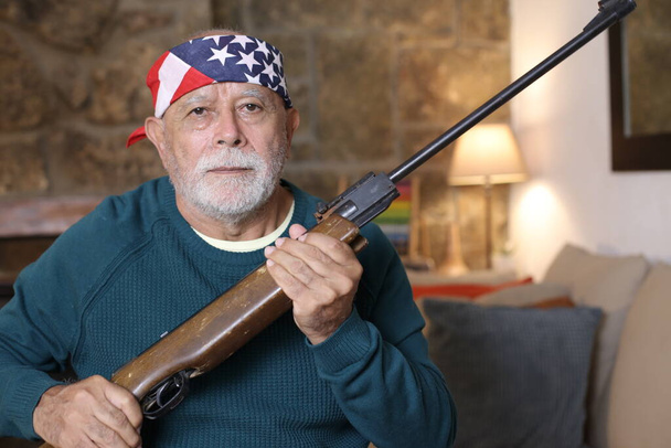 Nahaufnahme Porträt eines gut aussehenden älteren Mannes mit US-Fahnenband und Gewehr zu Hause - Foto, Bild