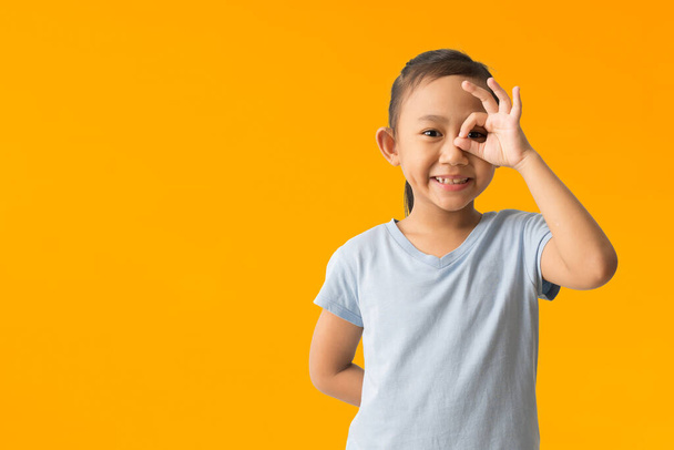 Happy asian child girl show OK sign over her eye, isolated on yellow background  - 写真・画像
