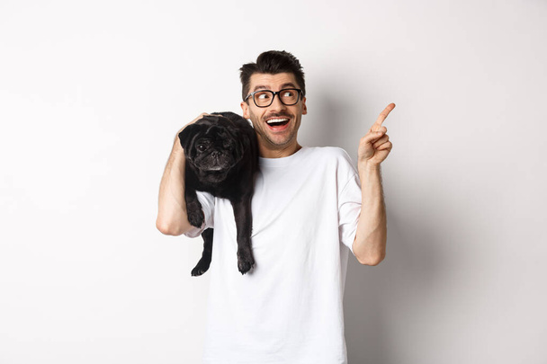 Εντυπωσιασμένος όμορφος άνδρας με γυαλιά, κρατώντας χαριτωμένο μαύρο σκυλάκι στον ώμο, δείχνοντας το δάχτυλο πάνω δεξιά γωνία στο λογότυπο του promo, στέκεται πάνω από το λευκό φόντο - Φωτογραφία, εικόνα