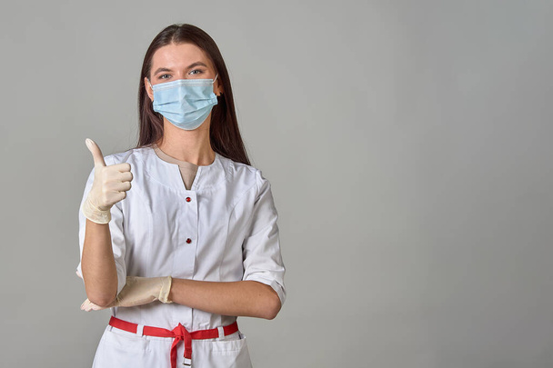 A doctor in a medical mask and a white coat shows his thumb on the background, copy space - Photo, Image