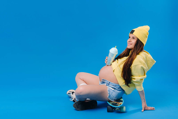 a pregnant girl in yellow clothes with a glass of juice sits on a skateboard on a blue background. - Фото, зображення