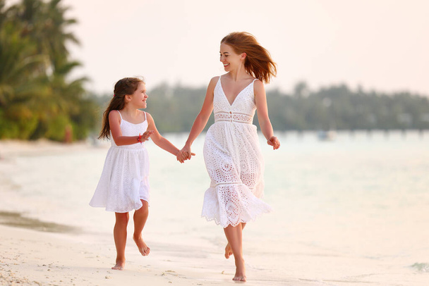 Optimistic family woman and girl in white dresses holding hands and looking at each other with smile while running on sandy beach near sea at sunny day - Foto, imagen