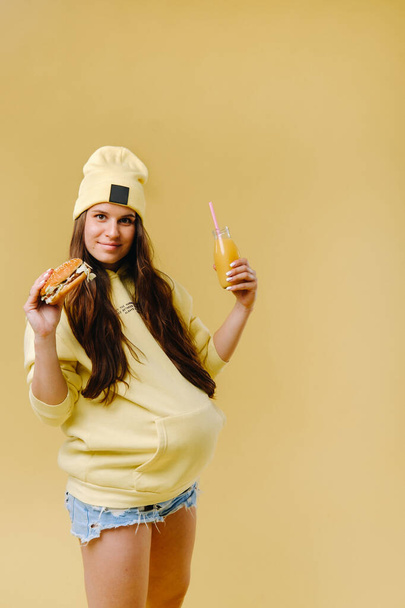 pregnant girl in yellow clothes with hamburgers in her hands on a yellow background. - Фото, изображение