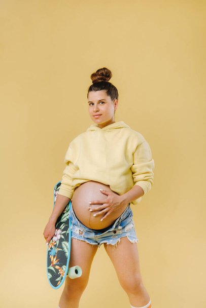 Pregnant girl in a yellow jacket with a skateboard in her hands on a yellow background. - Photo, Image