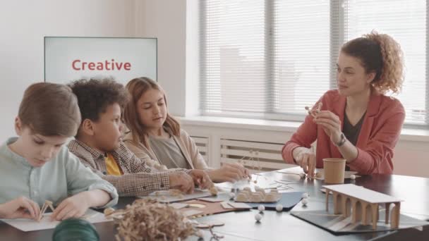 Klatka piersiowa młodej białej kobiety uczącej trójkę wieloetnicznych dzieci w wieku podstawówki, robiących rzemiosło na warsztatach, siedzących przy stole - Materiał filmowy, wideo