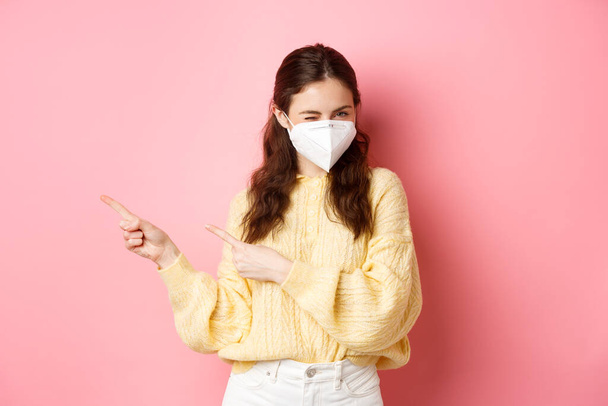 Covid, corona and social distancing concept. Coquettish young woman inviting to check out special offer, winking at you in medical respirator, pointing fingers aside at promo text, pink background - Fotografie, Obrázek