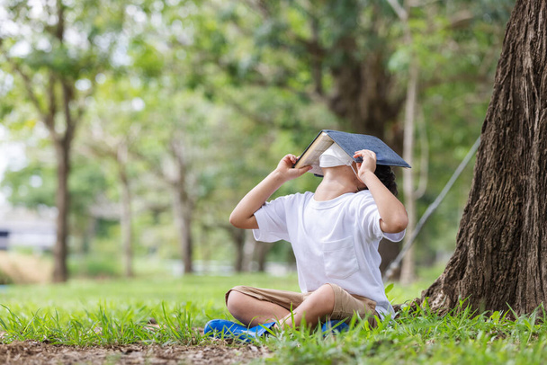 Valkoinen poika hygieenisessä naamiossa istuu yksin puun alla lukemassa kirjaa, joka peittää hänen päänsä.. - Valokuva, kuva