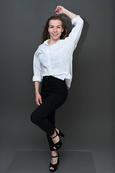 Photo in full growth of a slender caucasian girl with a smile and fluttering hair posing on a gray background in a white shirt and black trousers - Foto, imagen