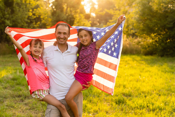 Szczęśliwa rodzina siedząca razem na podwórku z amerykańską flagą za sobą. Uśmiechnięta para z dziećmi świętuje amerykański dzień niepodległości trzymając amerykańską flagę - Zdjęcie, obraz