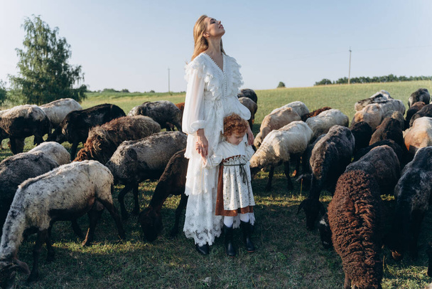 The beautiful woman in a white lace dress spending time with her little redhead daughter on the field with goats and sheep - Fotoğraf, Görsel
