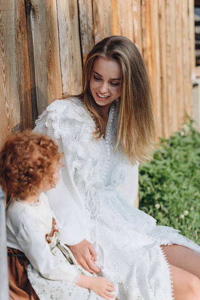 The beautiful woman in a white lace dress spending time with her little redhead daughter and sitting on the farm bench - Zdjęcie, obraz