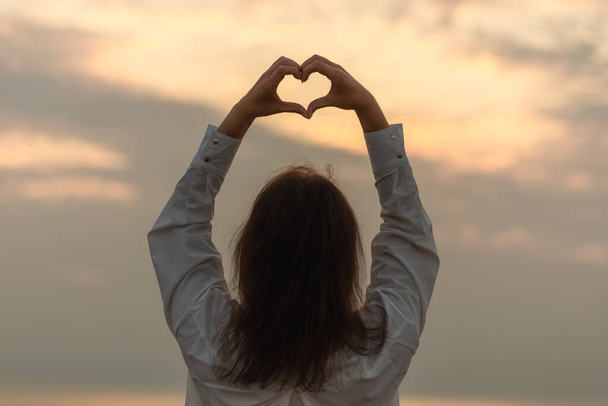 Young Woman holding a heart shape on the ocean, sea shore sunset eveningyoung beautiful woman makes heart by her hands at sunset summer,sea.Holiday concept.Back view. - Photo, Image