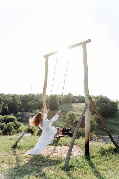 Back view of the beautiful woman in a white lace dress swinging on a wooden swing - Foto, Bild
