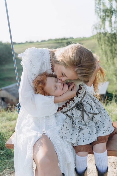 The beautiful woman in a white lace dress spending time with her little redhead daughter sitting on a swing - Photo, image