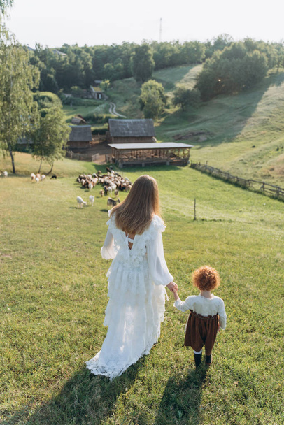 The beautiful woman in a white lace dress spending time with her little redhead daughter on the field with goats and sheep - Zdjęcie, obraz