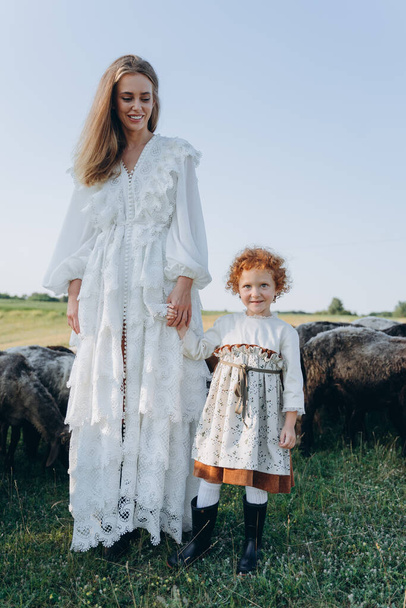 The beautiful woman in a white lace dress spending time with her little redhead daughter on the field with goats and sheep - Фото, изображение