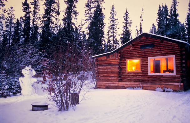 Άνετο κούτσουρο καμπίνα πλήρως φωτίζεται σε χιονισμένο δάσος χειμώνα με χιονάνθρωπο στο ρολόι μπροστά από το σπίτι διακοπών - Φωτογραφία, εικόνα