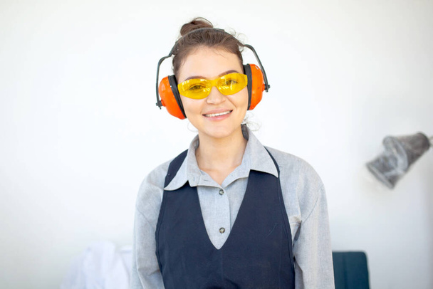 Joven mujer morena en gafas guantes y auriculares sonriendo lista para trabajar en taller de construcción - Foto, imagen