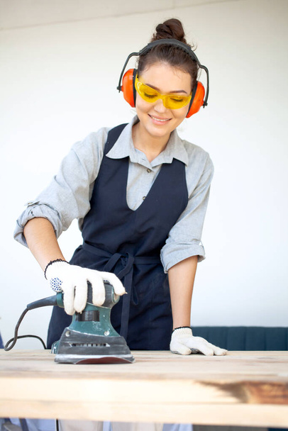 Mujer joven de cabello oscuro en gafas y auriculares sonriendo herramientas eléctricas en el taller preparando la superficie de la mesa de madera para pintar - Foto, imagen