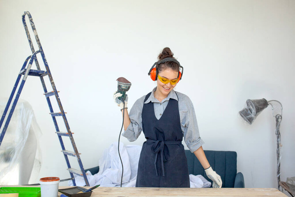 Mladá tmavovlasá žena v brýlích a sluchátkách s úsměvem běží elektrické nářadí v dílně připravuje dřevěný stůl na malování - Fotografie, Obrázek