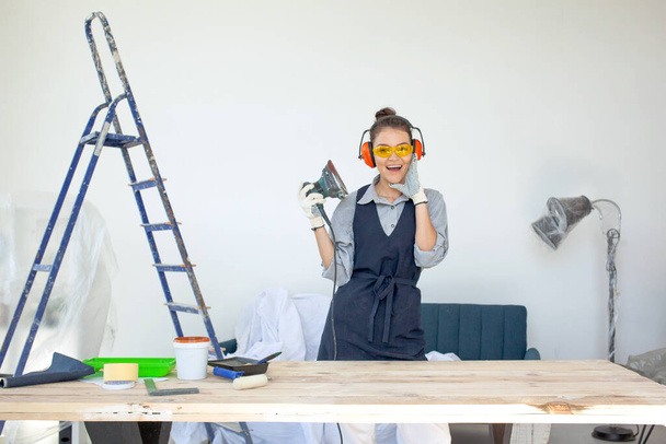 junge dunkelhaarige Frau mit Brille und Kopfhörer lächelnd Elektrowerkzeuge in der Werkstatt zur Vorbereitung der hölzernen Tischoberfläche für die Malerei - Foto, Bild