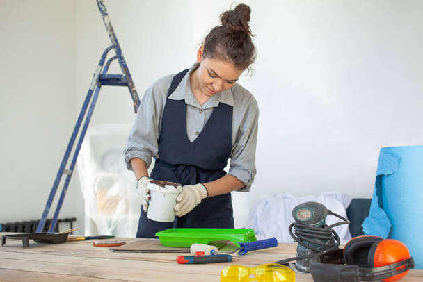 Joven mujer morena con guantes sonriente revolviendo y vierte pintura en el interior - Foto, Imagen